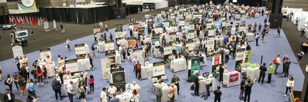 Aerial view of IFT FIRST Startup Pavilion with numerous startups exhibiting at their kiosks and attendees trying samples.