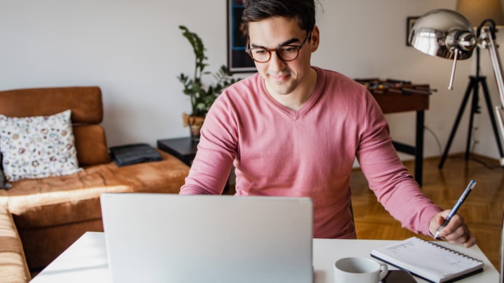 A young man with a laptop at home, he is working and writing notes in a notebook