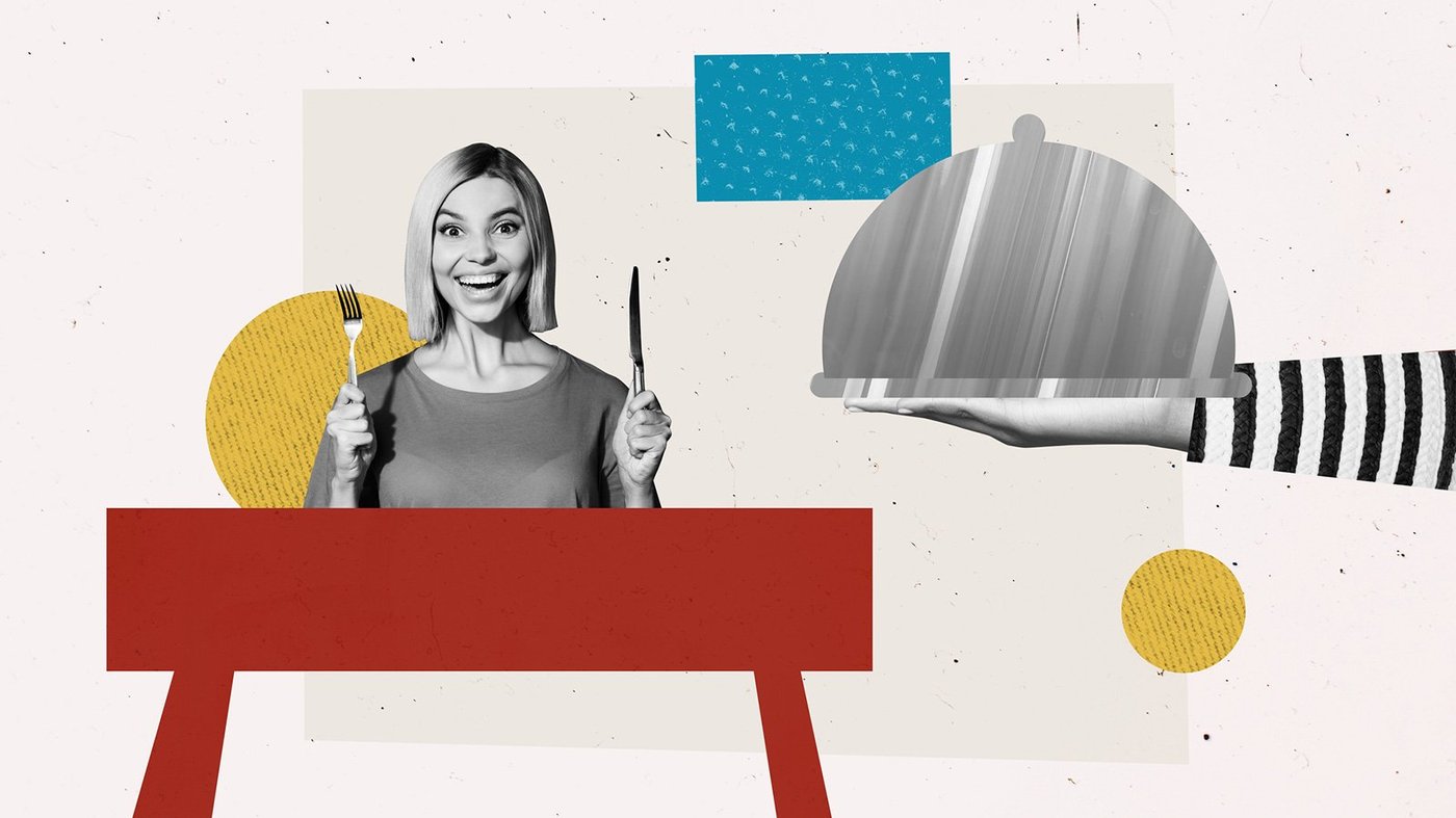 Excited woman holding a fork and knife ready to be served a plate of food with a cover on top.