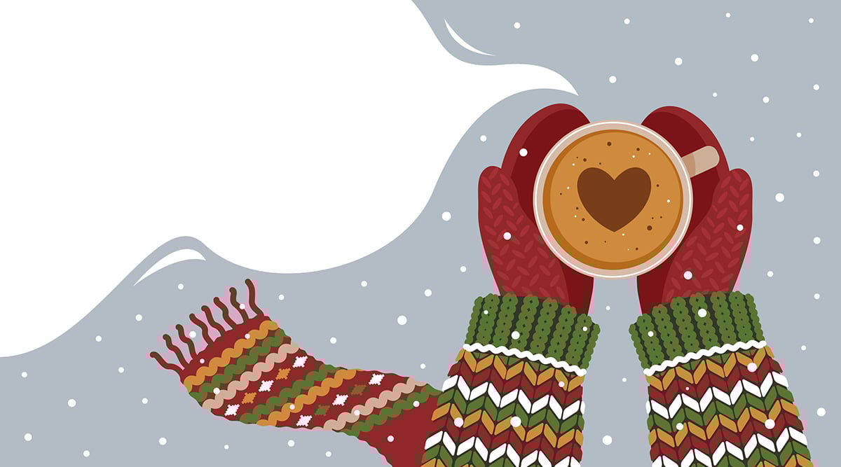 Person wearing a holiday sweater, scarf, and mittens holding hot cocoa with a heart out in the snow.