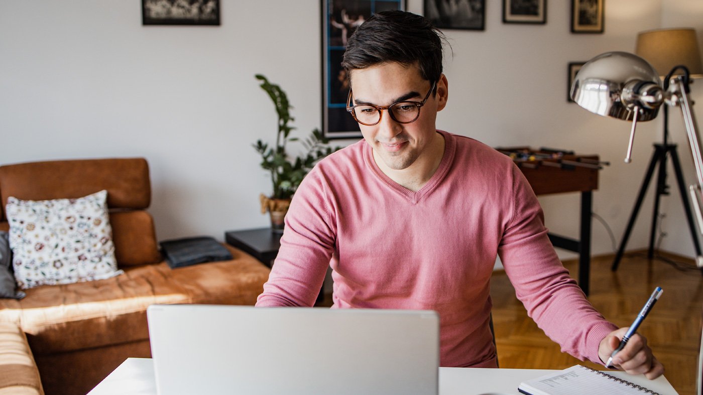 A young man with a laptop at home, he is working and writing notes in a notebook