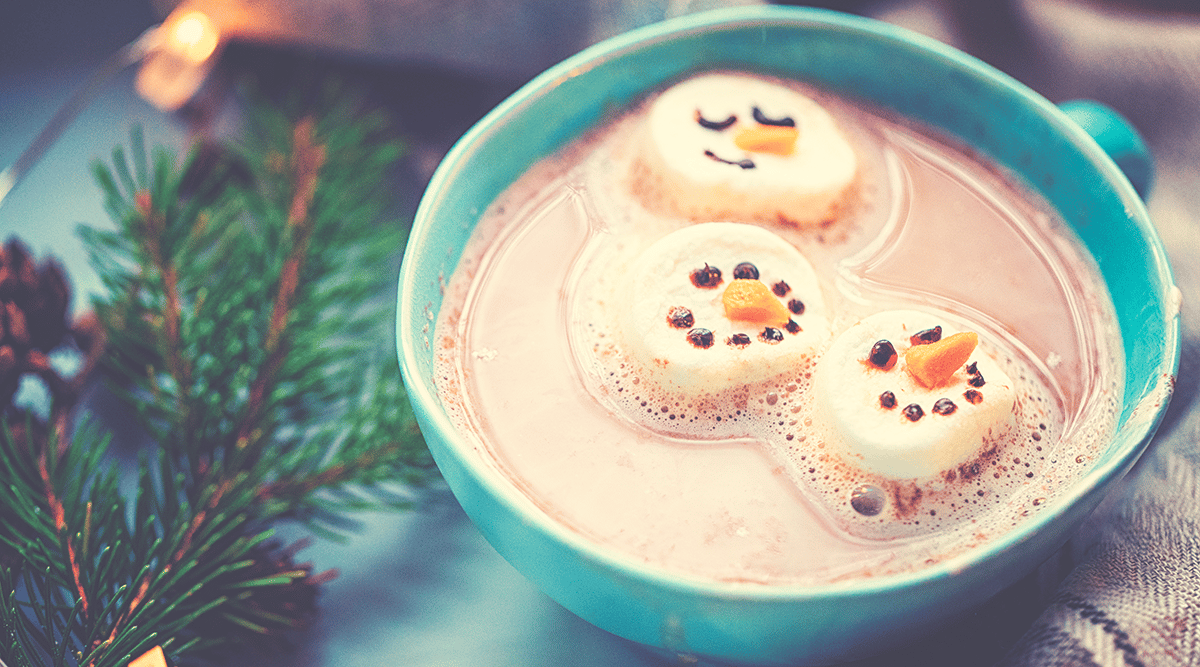 Hot cocoa with snowmen faces on marshmallows in a blue mug next to a pine branch