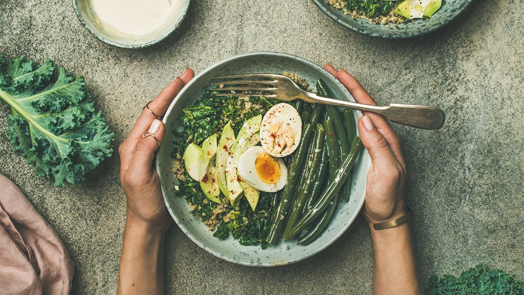Hands on a bowl filled with healthy food such as eggs, green beans, avocado, quinoa, and greens.