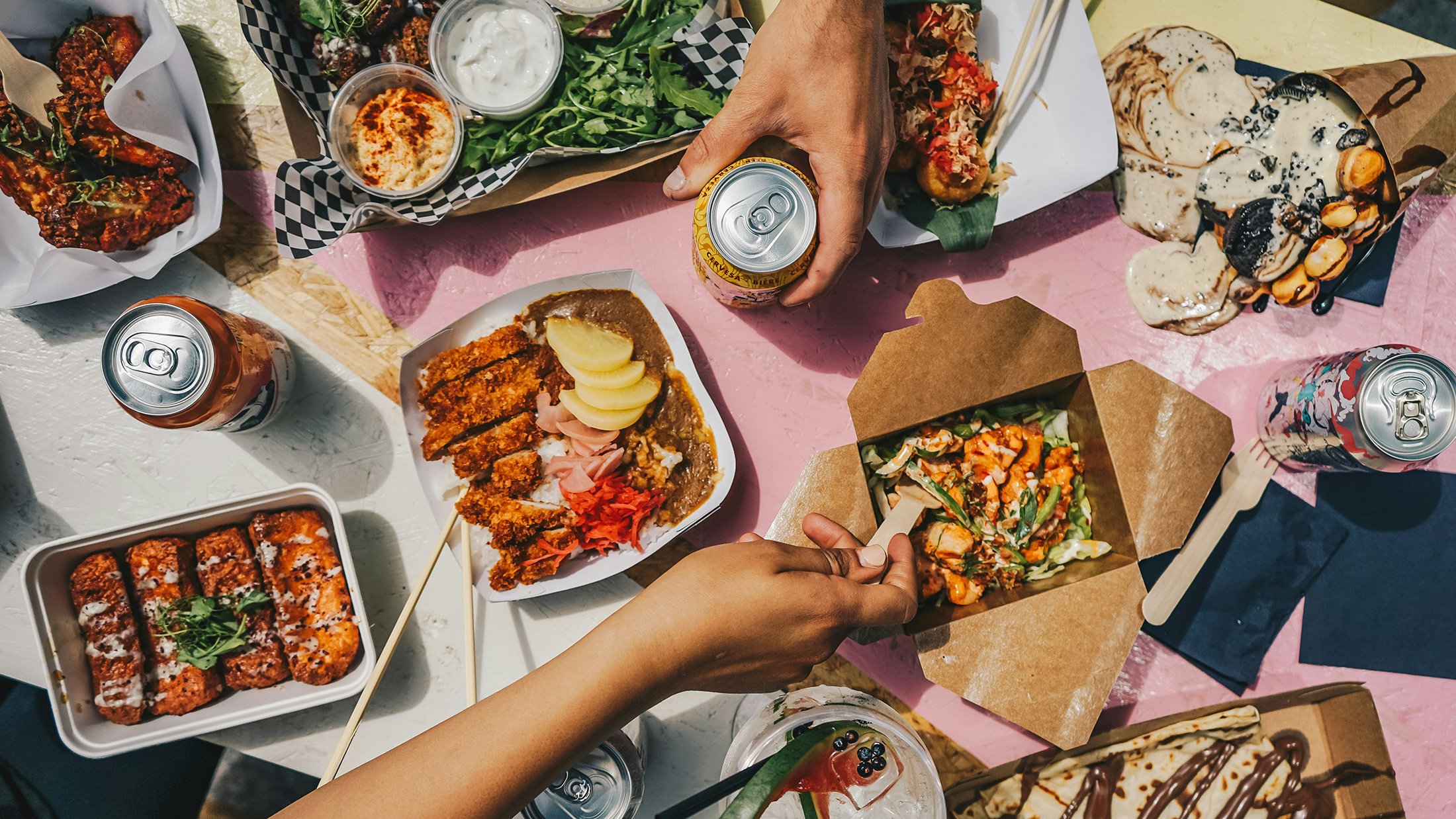 A feast of takeout food on a table with two different hands grabbing food and drink.