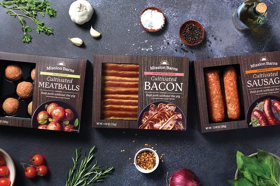 Packages of Mission Barns Cultivated Meatballs, Bacon, and Sausages on table surrounded by fresh ingredients