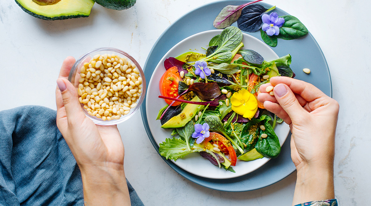 Hands adding pine nuts to a colorful salad.