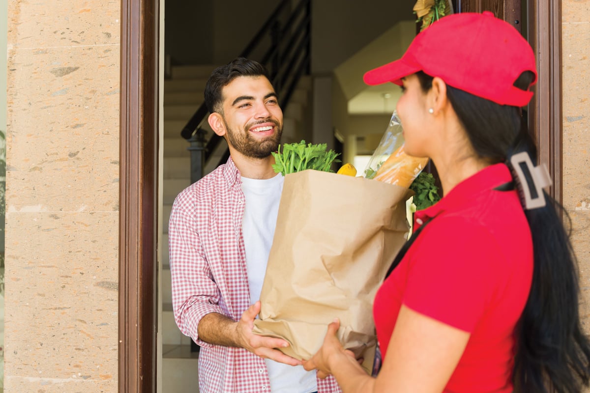 Delivery woman wearing a red uniform delivering a grocery bag to a young man at his front door.