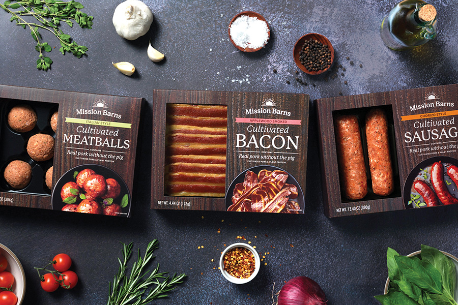 Packages of Mission Barns Cultivated Meatballs, Bacon, and Sausages on table surrounded by fresh ingredients